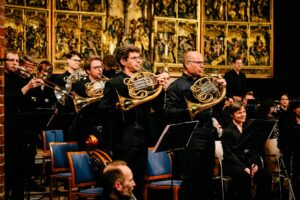 Horn spielende Personen in einem Orchester, im Hintergrund ein goldener Hochaltar