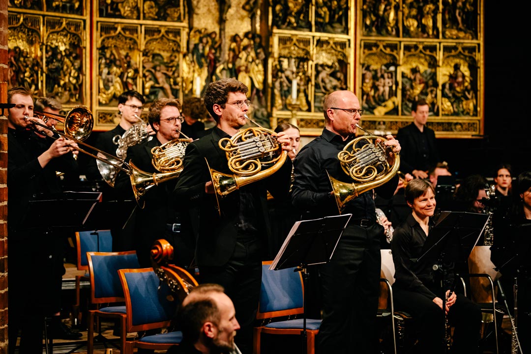 Horn spielende Personen in einem Orchester, im Hintergrund ein goldener Hochaltar