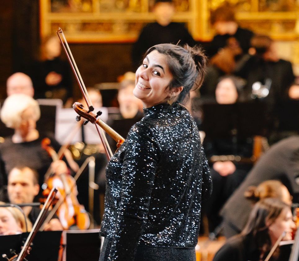 Eine Frau steht mit einer Geige im Arm vor einem Orchester